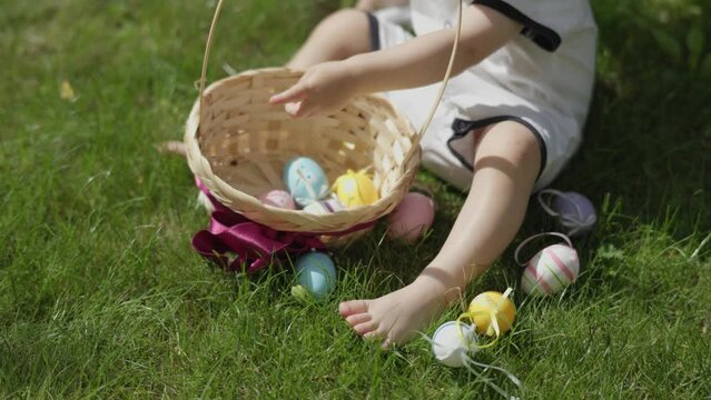 close-up toddler baby hands put colorful Easter eggs into basket in green grass lawn during warm sunny spring day. unrecognizable child sit on green grass play Easter eggs after traditional game