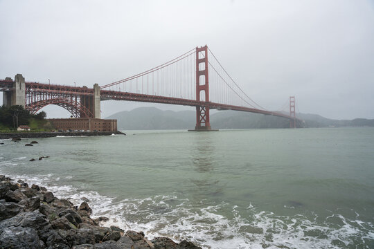 Dramatic image of the Golden Gate Bridge during a rainstorm with a rocky beach in the foreground.