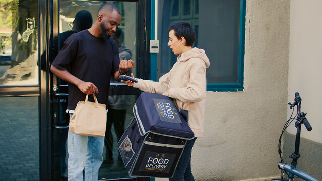 Young client taking restaurant meal order using smartwatch to pay with nfc on pos, paying for takeaway package. Food delivery service courier giving fastfood in paperbag, contactless payment.