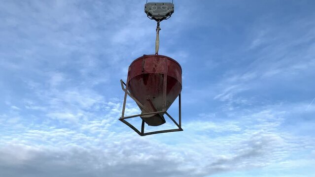 Red bucket with concrete hung from crane in the middle of a blue sky. Tilt up shot of a bucket of cement holded by a hook with a face painted in it. Steel cement container hanging in the air.
