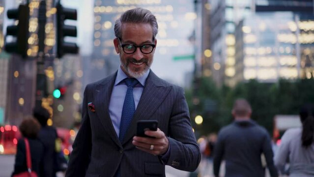 Handsome and Successful business man in stylish suit looking  at smartphone screen while walks outdoors in city. Prosperous smiling entrepreneur in eye glasses feels confident and prosperous