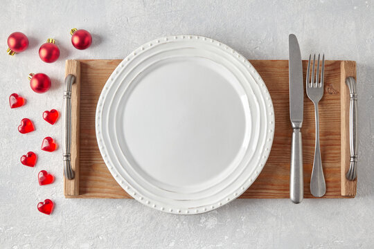 An Empty White Ceramic Plate With Cutlery On A Wooden Tray Surrounded By Red Christmas Balls And Hearts On A Light Concrete Table