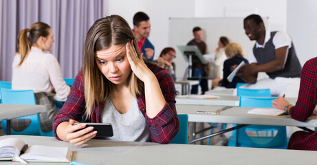 Portrait of upset girl student sitting apart in auditorium, looking at phone