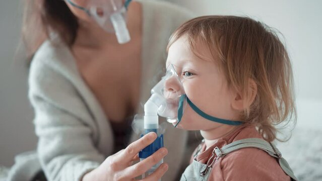 Asian mother helping sick daughter use nebulizer while embracing her on couch at home. Woman makes inhalation with equipment to toddler boy. Ill child lying on couch having respiratory illness helped