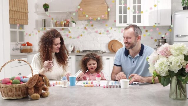 Portrait of lovely family decorating traditional Easter eggs with their cute, little daughter. Laughing, happy mother and father helping their girl to paint Easter symbols. High quality 4k footage
