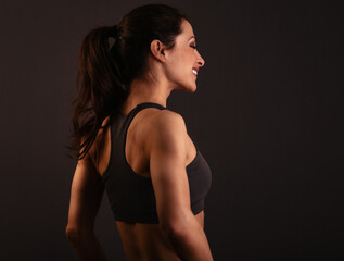 Naklejka premium Smiling happy female sporty muscular with ponytail with stretching workout of shoulders, blades and arms in sport bra, standing on dark grey background with empty copy space. Back view.