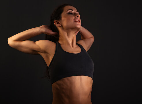 Happy smiling sporty muscular woman doing stretching workout on her spine in the cervical and thoracic regions and shoulders in sport bra, holding the hands in lock behind the head on grey background