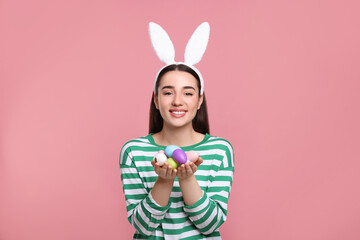 Happy woman in bunny ears headband holding painted Easter eggs on pink background