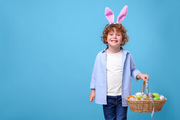 Happy boy in cute bunny ears headband holding wicker basket with Easter eggs on light blue...