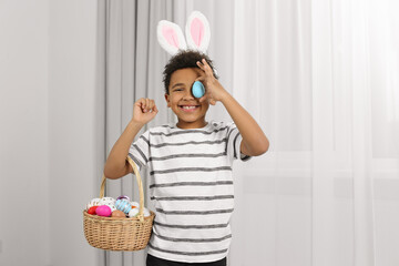 Cute African American boy in bunny ears headband covering eye with Easter egg indoors
