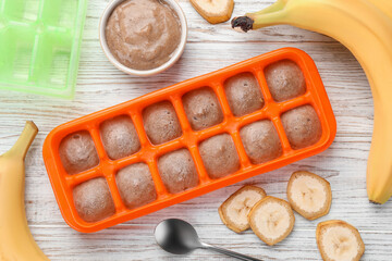 Banana puree in ice cube tray with ingredients on white wooden table, flat lay