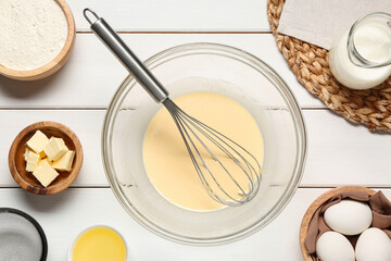 Glass bowl of crepe batter with whisk and ingredients on white wooden table, flat lay