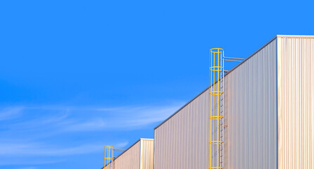 Fototapeta na wymiar Perspective side view of 2 factory buildings with yellow cylinder ladder on aluminium corrugated wall against blue sky background, widescreen view with copy space 
