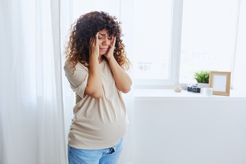 A pregnant woman stands in the house at the window, tired and has a headache, tears, crying from fatigue in a home T-shirt, the complexity of motherhood