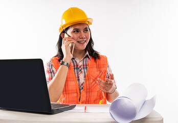 a female architect in safety waistcoat sitting in front of the opened laptop while calling someone on phone on isolated background