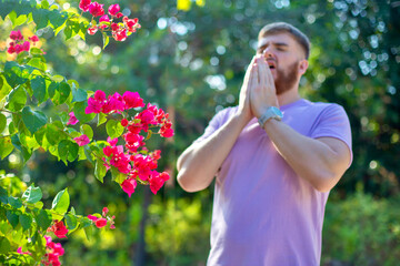 Fototapeta premium Portrait of handsome young allergic man is suffering from pollen allergy or cold on natural flowers, flowering tree background at spring or sunny summer day, sneezes, blowing his runny nose rubs eyes