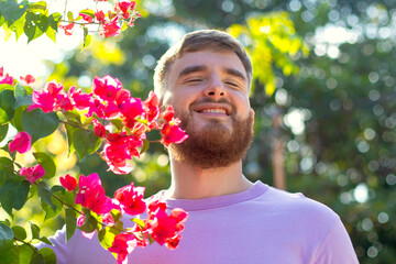 Portrait of happy handsome bearded guy, young positive man with beard is smelling beautiful red pink flowers in the garden, smiling, enjoying spring or summer day, breathing deep deeply fresh air - obrazy, fototapety, plakaty