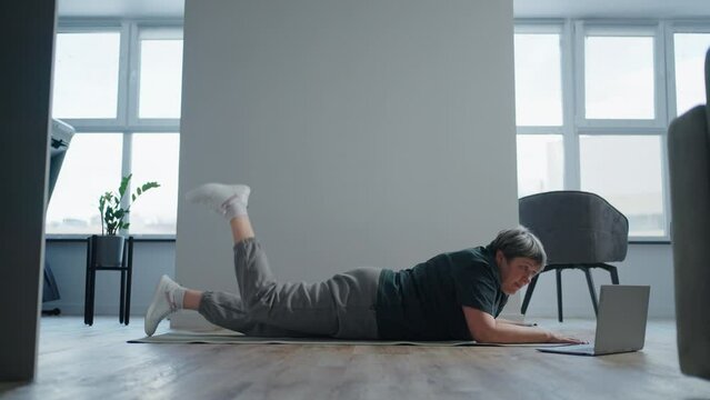 senior woman watching workout online by laptop and training at home, lying on floor and lifting leg