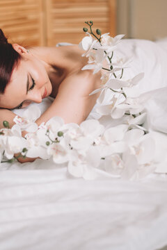 Photo shoot for women's day. The girl lies on the sheet. Naked female body in a sheet. Girl and orchids. Girl with flowers.