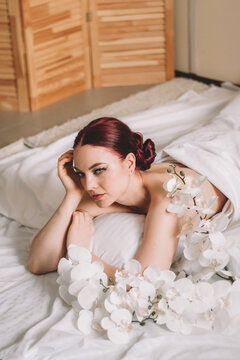 Photo shoot for women's day. The girl lies on the sheet. Naked female body in a sheet. Girl and orchids. Girl with flowers.