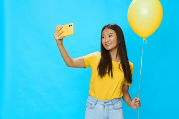 Asian woman holding the phone and looking at the screen talking on a video call with a yellow balloon in her hands on a blue background in a yellow T-shirt smiling with teeth 