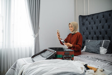 beautiful woman in hijab holding and using cell phone while preparing suitcase in bedroom