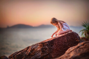 Little girl with long blond hair on the ocean. Baby on the seashore. Beautiful girl looks at the ocean. The girl is sitting on a big stone. Sea waves. Baby at sunset.