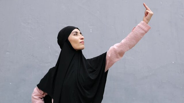 Islamic Muslim woman in black hijab and pink shirt stands in confident posture and extends her hand to sky, showing the middle finger up. Blasphemy, sinfulness, crime against religion, abusive gesture