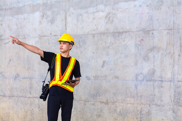 Asian engineer wearing hard hat and safety vest pointing finger while standing holding tablet and hang up camera in construction site.Copy space