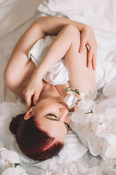 Photo shoot for women's day. The girl lies on the sheet. Naked female body in a sheet. Girl and orchids. Girl with flowers.
