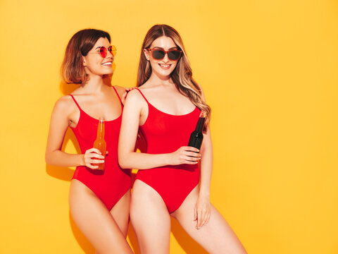 Two beautiful sexy smiling hipster women in summer swimwear bathing suits. Trendy models in studio. Hot female isolated on yellow. In sunglasses, holding bottle of lemonade, drinking