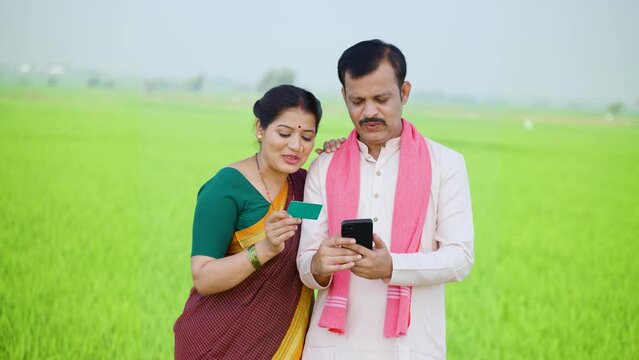 Happy smiling village couple farmer making online payment using credit card on mobile phone at farmland - concept of growth, banking or financial and ecommerce shopping.