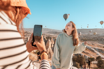 Two happy girl friends taking photos and video on phone for social networks in Cappadocia, Turkey. Travel and influencer concept - obrazy, fototapety, plakaty
