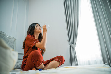 asian girl in pajamas wakes up sitting in bed drinking water from a bottle in the morning
