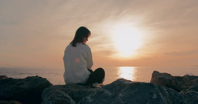 Woman with alley cat watching the sunset on rocky sea coast