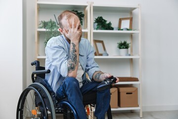 A man in a wheelchair gamer plays games with a joystick in his hands at home, anger and sadness from losing, copy space, with tattoos on his hands, health concept man with disabilities, real person