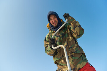 A warmly dressed Asian guy drills a hole in the ice on winter fi