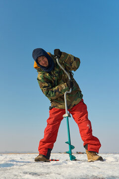 A warmly dressed Asian guy drills a hole in the ice on winter fi