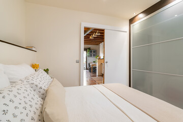 Cozy small bedroom in white tones with double bed glass wardrobe and shelf with open door overlooking the kitchen. Concept of cozy housing for a small family