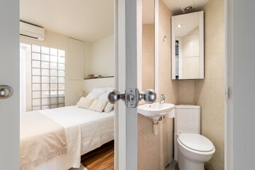 View from bathroom with toilet with beige tiles toilet bowl and sink to bedroom with bed and glass brick wall. Concept of cramped but thoughtful interiors and space