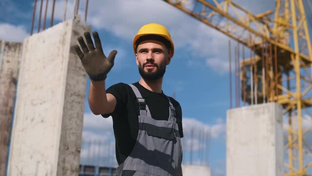Showing stop gesture by hand. Young engineer in hard hat and uniform is on the construction site.
