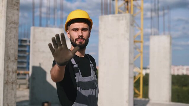 Showing stop gesture by hand. Young engineer in hard hat and uniform is on the construction site.