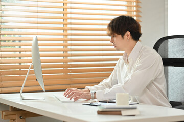 Elegant young businessman in white shirt looking at computer monitor, reviewing project in bright office interior