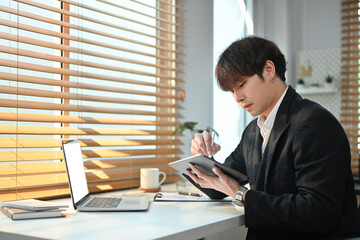Image of focused young businessman using digital tablet, reviewing project in bright office interior