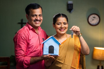 Happy smiling couple showing new house keys by toy home while looking camera - concept of new home...