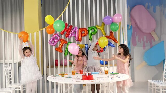 Cheerful children are playing at a birthday party, they throw balloons and spin around in a brightly decorated room. There is a delicious red cake on the table.