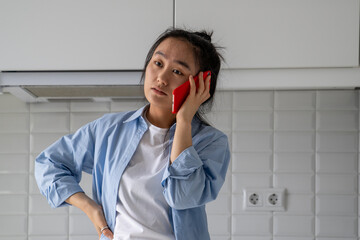 Upset dissatisfied Asian woman talking on phone arguing by mobile while standing in kitchen at...