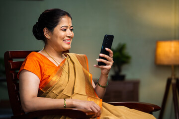Middle woman in sare buy using mobile phone on chair at home - concept of technology, social media sharing, internet.