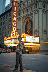 Portrait of a long-haired blonde young girl with a sweatshirt, backpack and vintage photo camera sightseeing at night in Chicago, in front of the famous theater