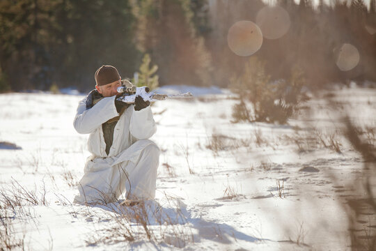 A hunter wearing  a cold-weather camouflage outfit aims a rifel while crouching in a snow-covered field.
